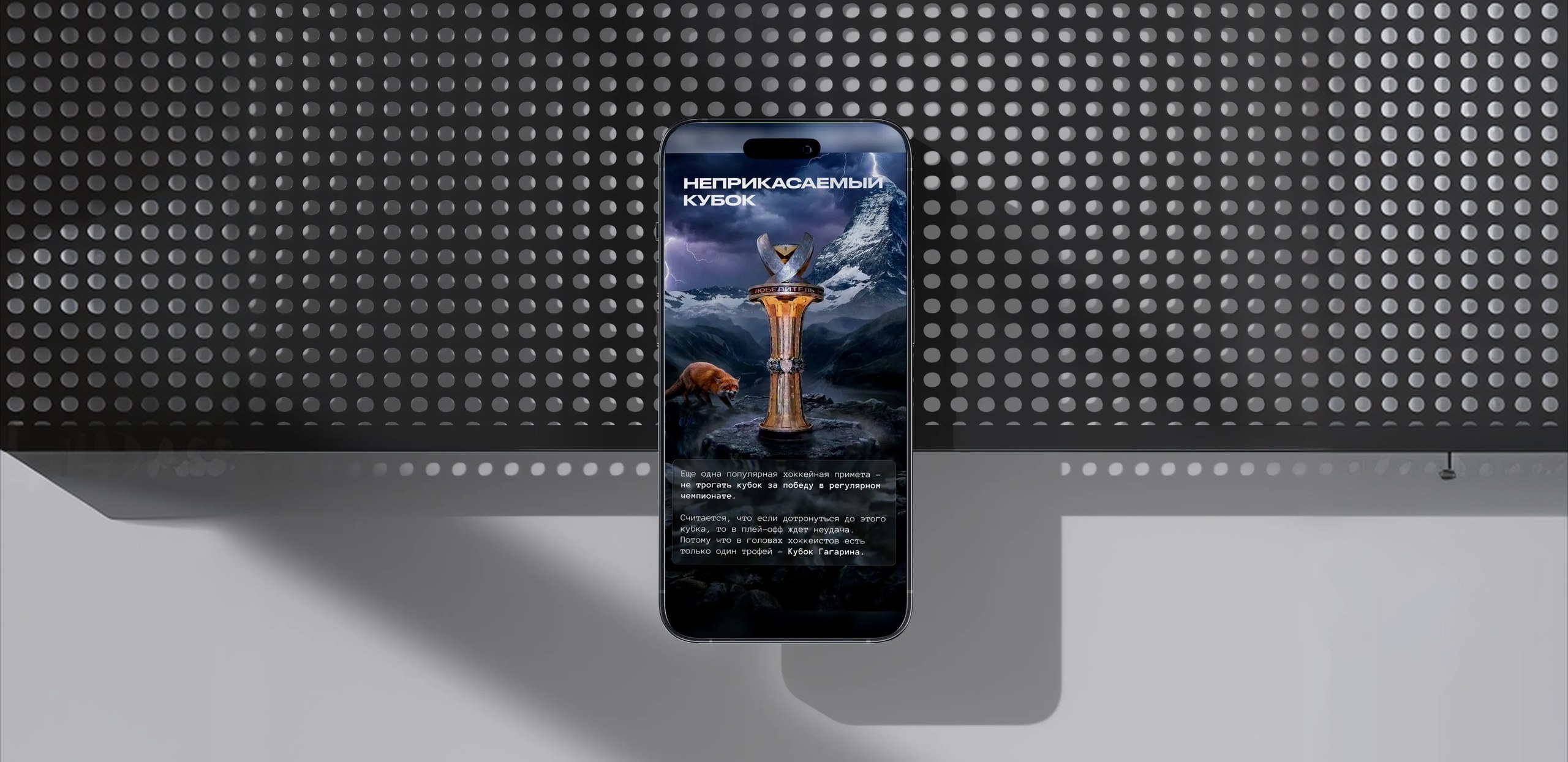 A phone on a dark perforated panel showing the "НЕПРИКАСАЕМЫЙ КУБОК" story